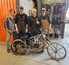 Four students stand wearing coveralls in a welding centre. In front of them sits a welded motorbike sculpture with skeletal accents.