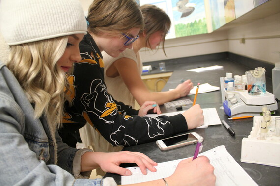 Three students in a science classroom stand at a counter littered with test tubes filled with solutions. There are also notebooks and other school supplies on the counter.