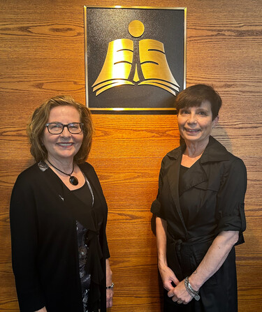 Two women stand next to a golden plaque of the Elk Island Public Schools logo hanging on a brown wooden wall.