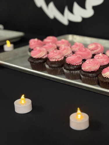 A tray of chocolate cupcakes with pink frosting sit on a black tablecloth surrounded by small tea lights.