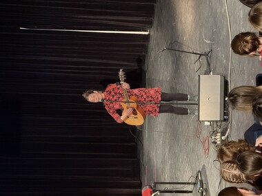 A person stands on stage playing a guitar and singing into a microphone, while students watch as they sit in a school auditorium.