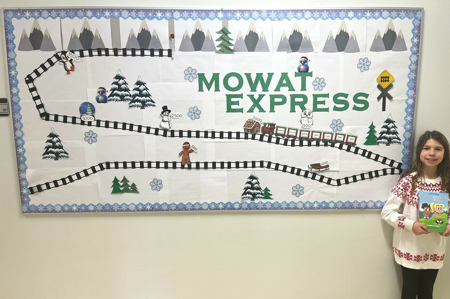 An elementary student stands on the right-hand side of the image holding a book. In the centre of the image is a bulletin board displaying a visual of the Mowat Express reading challenge, fit with paper railway track and train amidst a snowy environment.