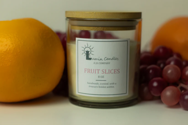 A candle sits in between an orange to the left and grapes nestled beside and behind the candle to the right. Further back behind the grapes is another orange. The off-white candle in the middle is inside of a clear glass container that has a wooden top an