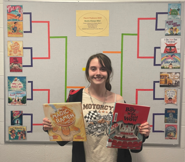 A student stands in front of a bulletin board with printed book covers set up in a tournament-style bracket, eight on each side. The student smiles at the camera while holding up two books.