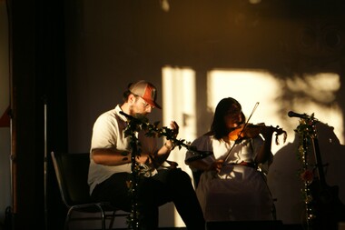 Two individuals sit in a dim room lit by a sunny window as they play fiddles together.