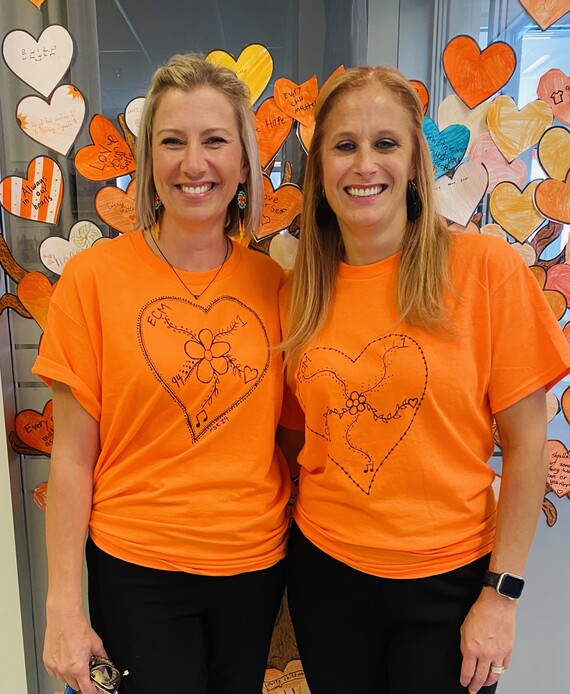 Two staff members stand together and smile at the camera while wearing orange shirts with a heart design and a flower in the middle of the design. In the background, there's a clear glass window with hearts of different shades of orange and varied pattern