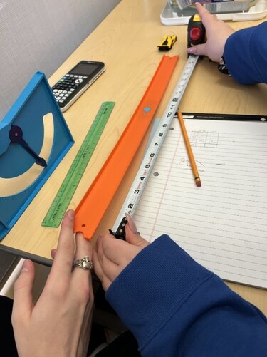 On a desk, there's a student's hands holding a ruler up to a Hot Wheels car slope being held by the hand of another student. Nearby is a notepad, pencil and ruler.