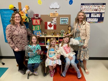 Two adults stand in between three students sitting down in chairs. The students hold books and stuffed animals. Behind them is a display filled with shelved books, building blocks, puppets and more age-appropriate resources for developing literacy skills.