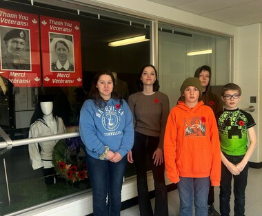 Five students stand beside a display behind glass in a school hallway. The students all wear poppies on their left-hand side above their hearts. To the left of the image, the display is visible, consisting of a uniform, wreath and two posters.