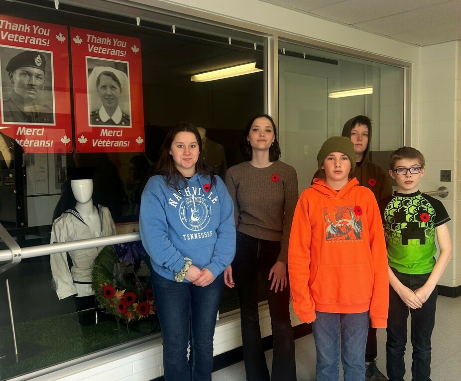 Five students stand beside a display behind glass in a school hallway. The students all wear poppies on their left-hand side above their hearts. To the left of the image, the display is visible, consisting of a uniform, wreath and two posters.