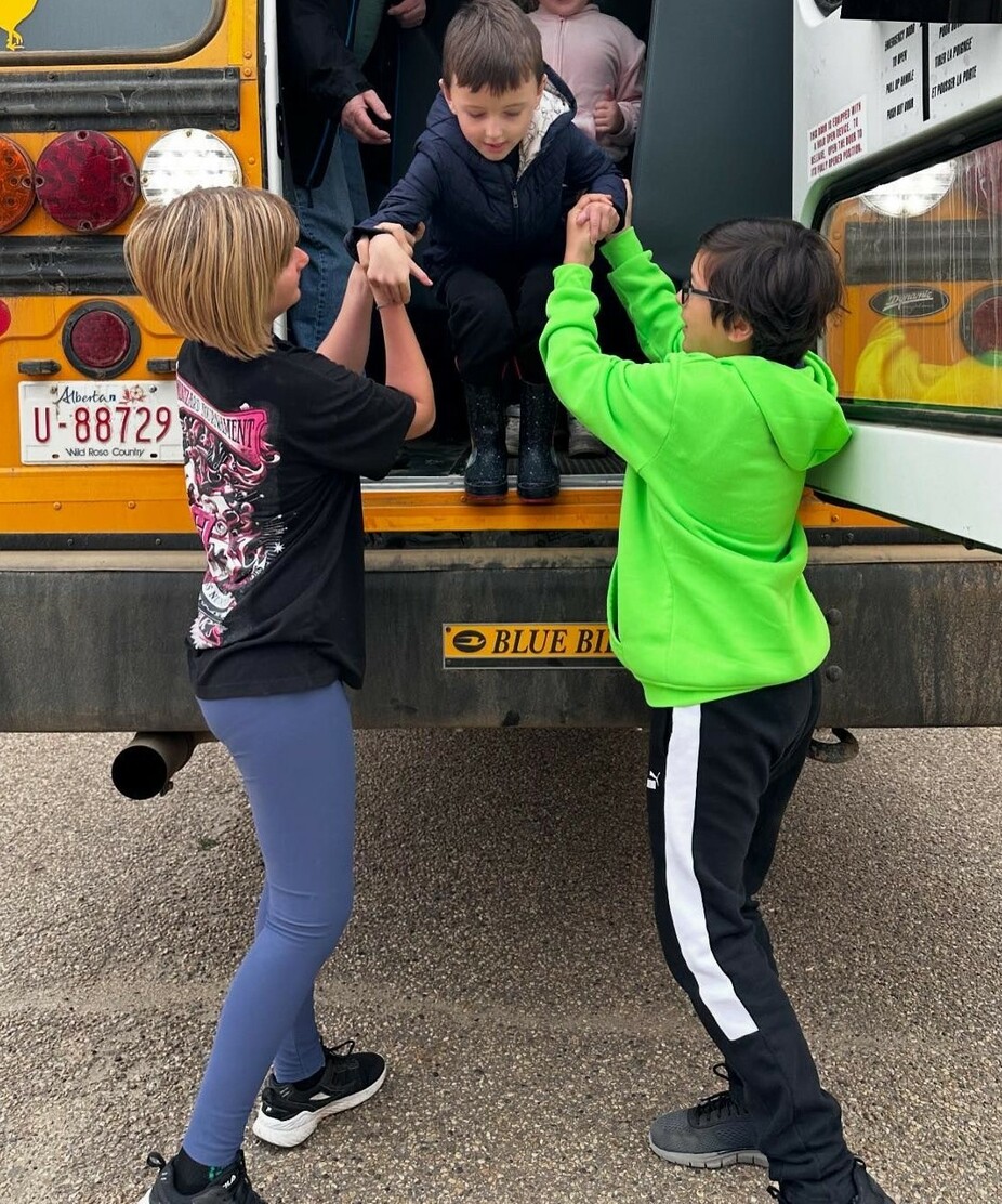 An adult and a student stand at the back of a bus holding the arms of another student as they help them down from the emergency exit.