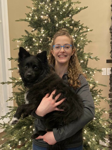 A woman stands in front of a Christmas tree decorated in white lights. She smiles at the camera and holds a black dog in her arms.