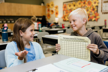 Two students sit at a desk as they smile and talk with one another. The student on the right-hand side of the image holds up a mathematics worksheet about decreasing sequences.