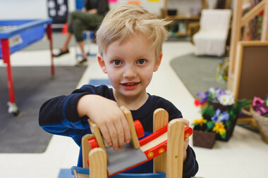 A prekindergarten student with short blonde hair looks at the camera as they play with a toy that has a slope and slide mechanism in a classroom.