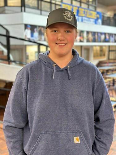 A male senior high student standing with his hands in his pockets wearing a blue hoodie and dark grey baseball cap smiling at the camera.