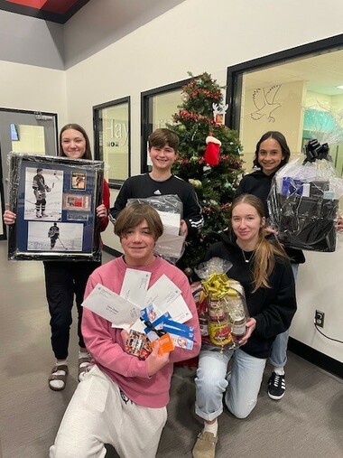 Five students gather around a Christmas tree each holding a silent auction item such as hockey memorabilia, gift baskets and gift cards.