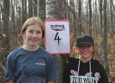 Two students stand beside a tree with a sign that has the text Zombie Run and a large number four beneath it.