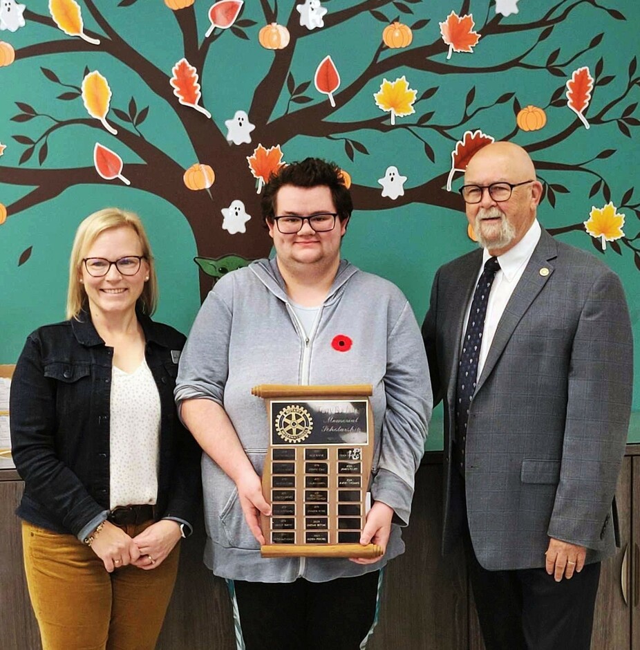 Three individuals stand in front of a bulletin board displaying a tree littered with autumn leaves. They all smile at the camera. The person in the middle holds a plaque filled with names of people who have won the Fort Sask Rotary Club Scholarship.